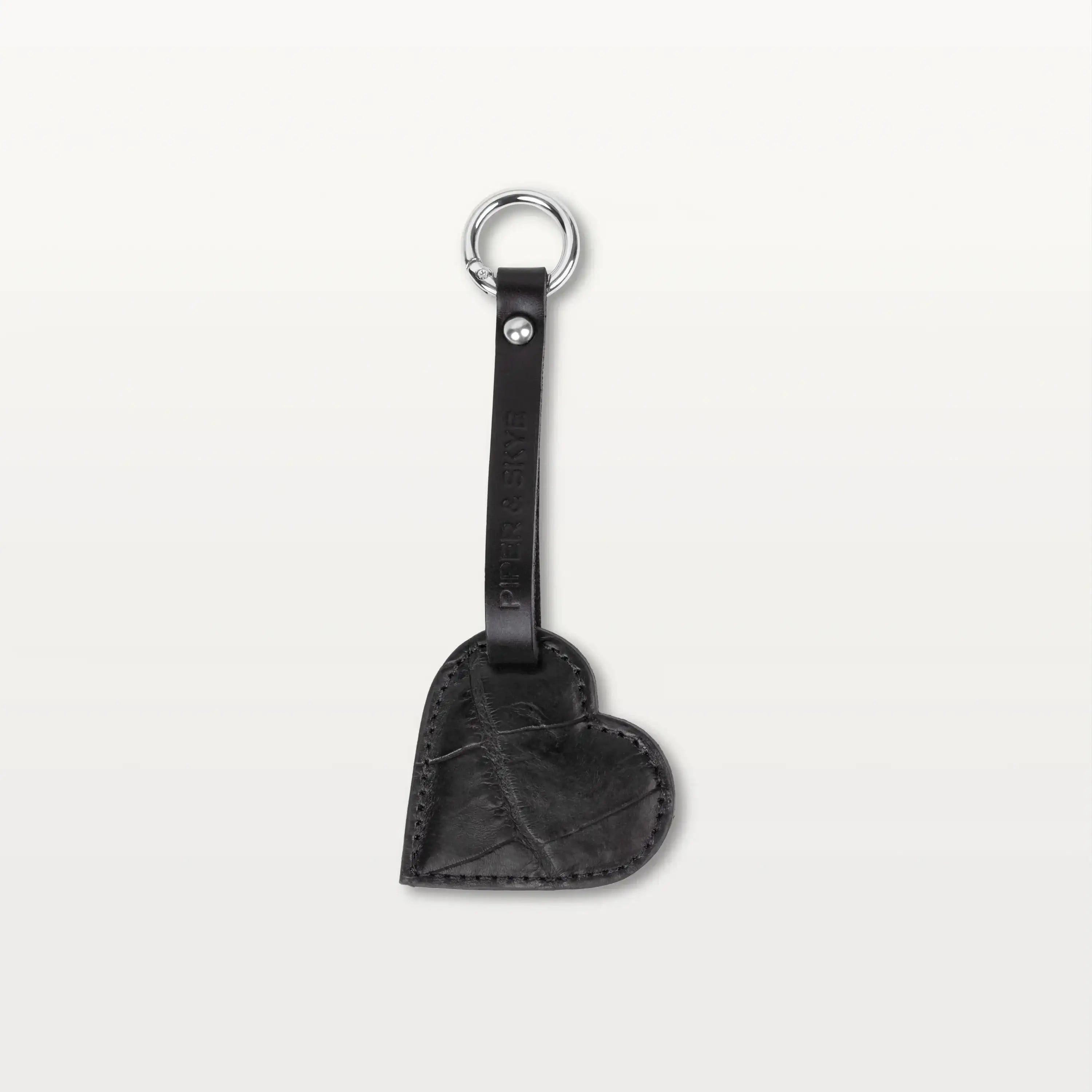 A heart-shaped keychain made from black alligator leather hanging from a thin leather strap with a silver metallic ring.