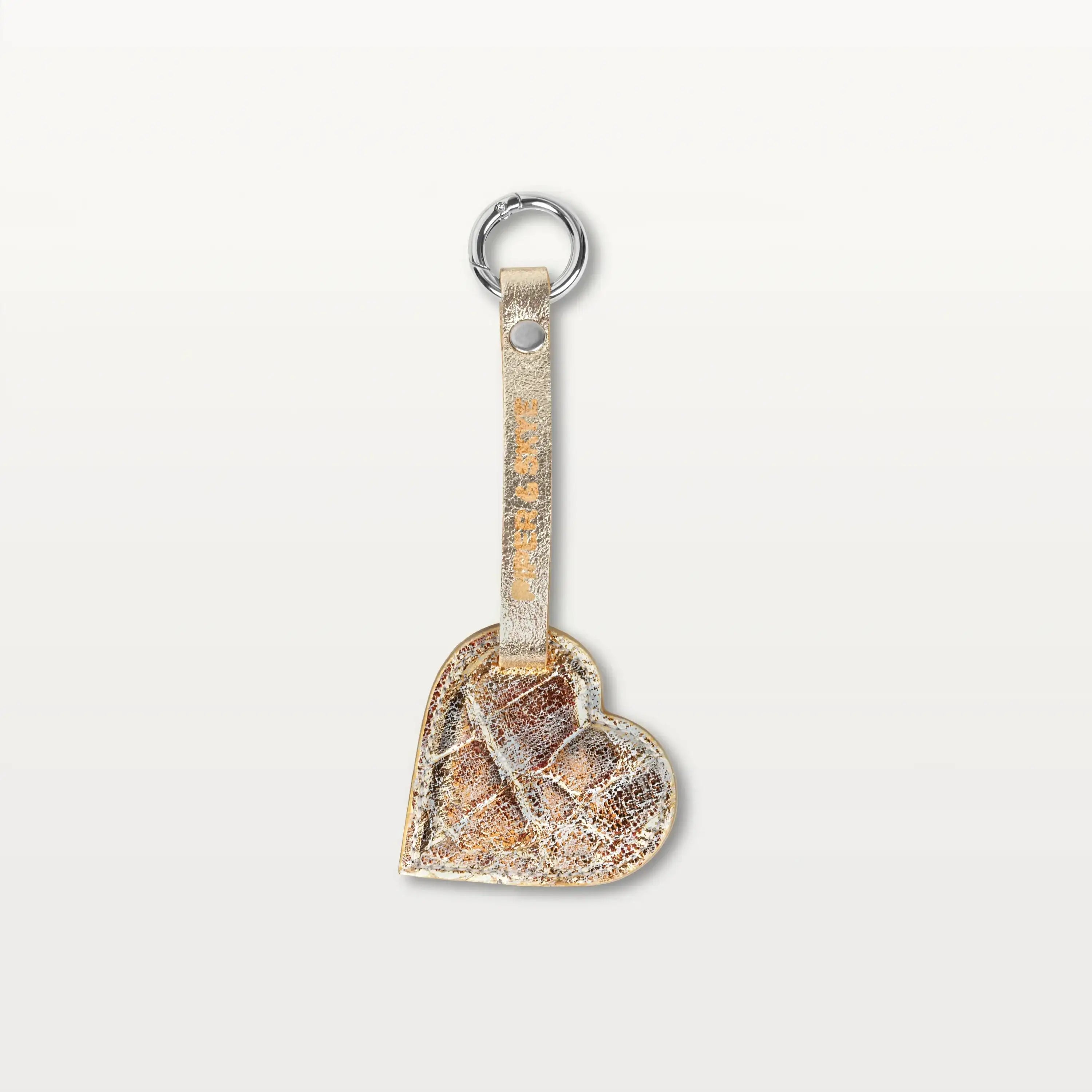 A heart-shaped keychain made from metallic gold alligator leather hanging from a thin leather strap with a silver metallic ring.