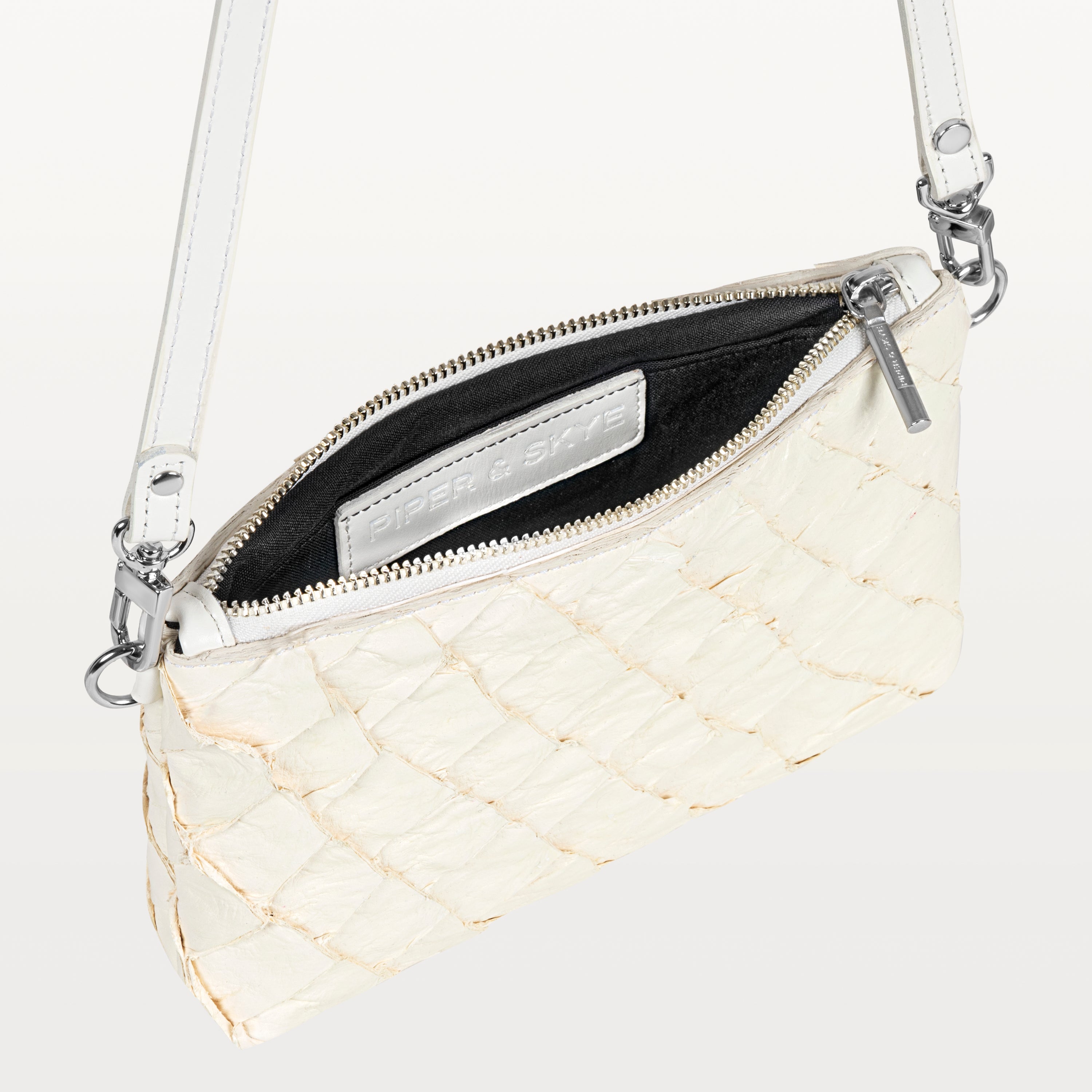 Small white crossbody bag made of pirarucu leather with its silver zipper open revealing the black interior and Piper & Skye logo. 
