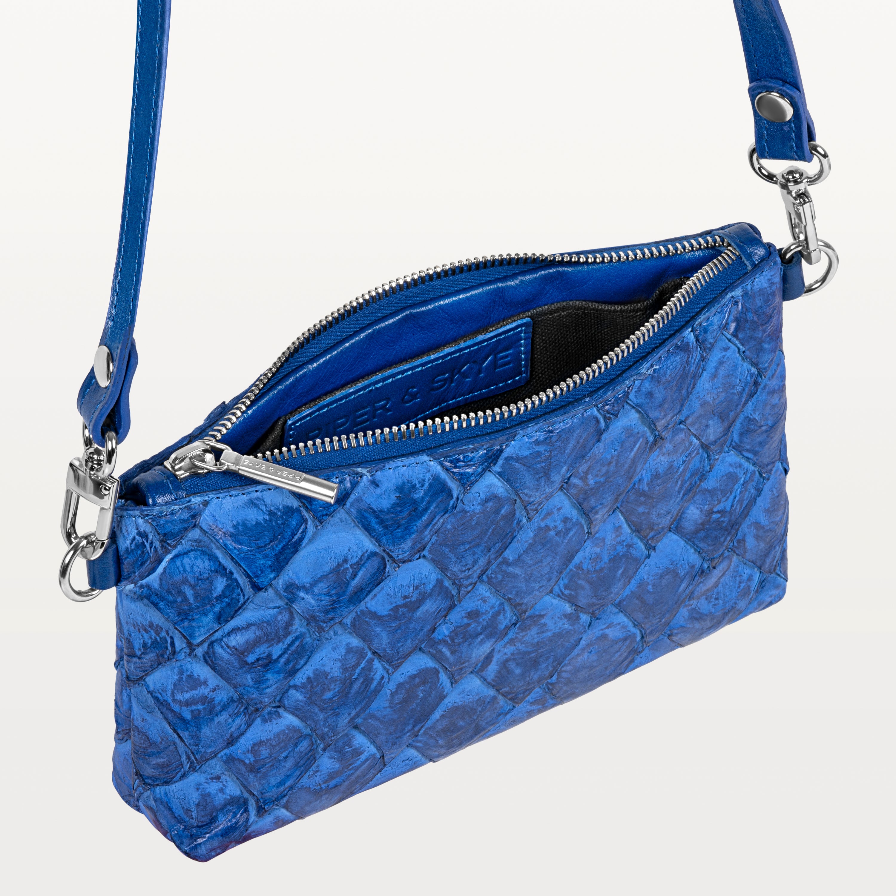 Small cobalt blue crossbody bag made of pirarucu leather with its silver top zipper open revealing the black interior and Piper & Skye logo. 