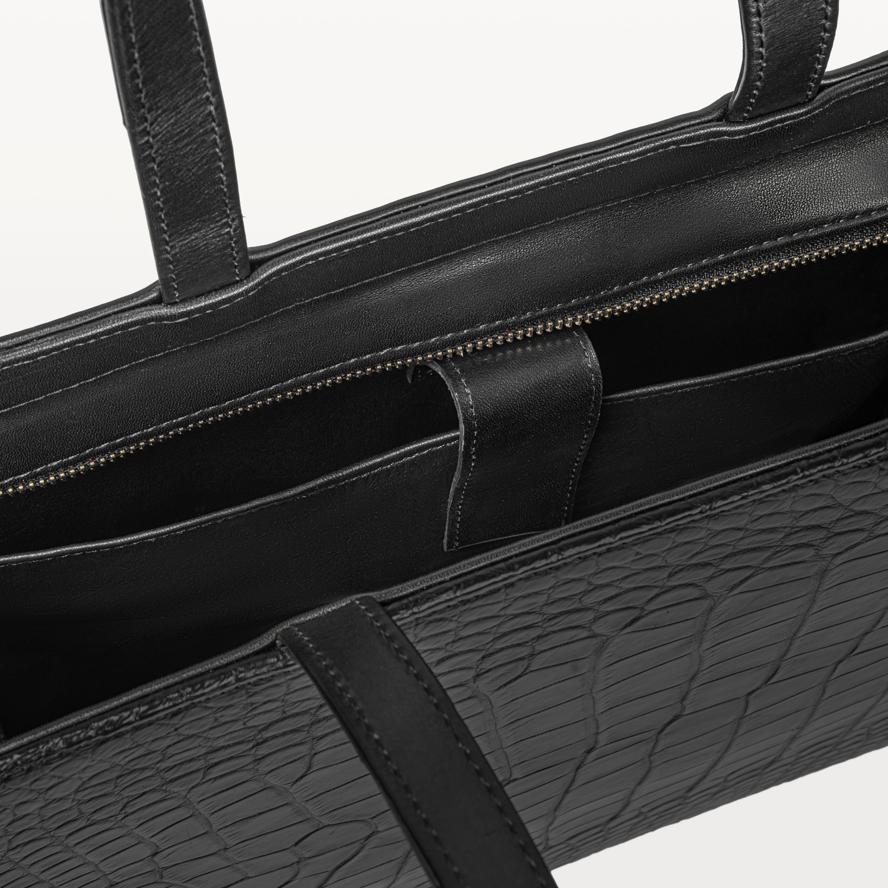 Top zipper of the Braemar tote bag in black alligator leather open to show the inner laptop sleeve. 