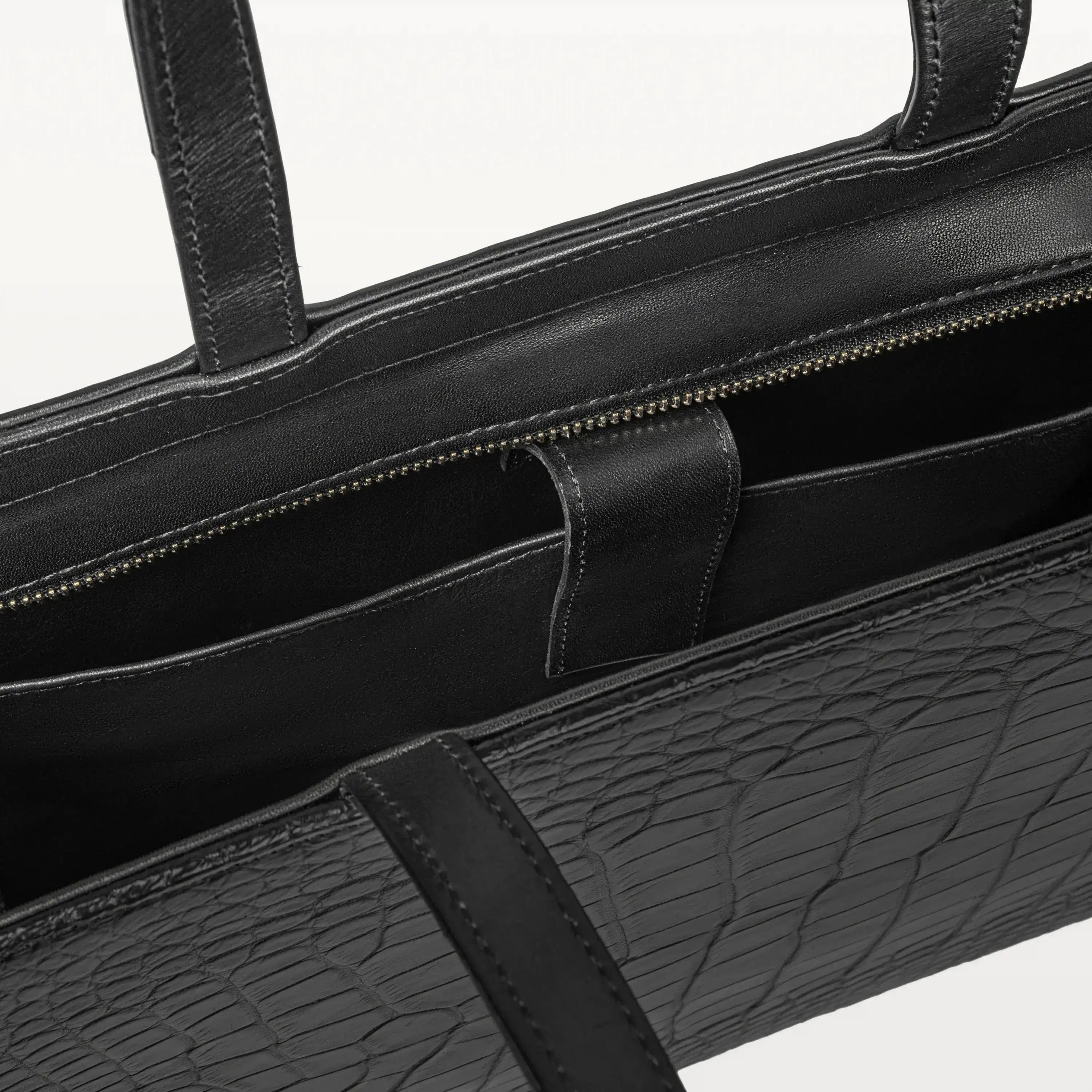 Top zipper of the Braemar tote bag in black alligator leather open to show the inner laptop sleeve. 