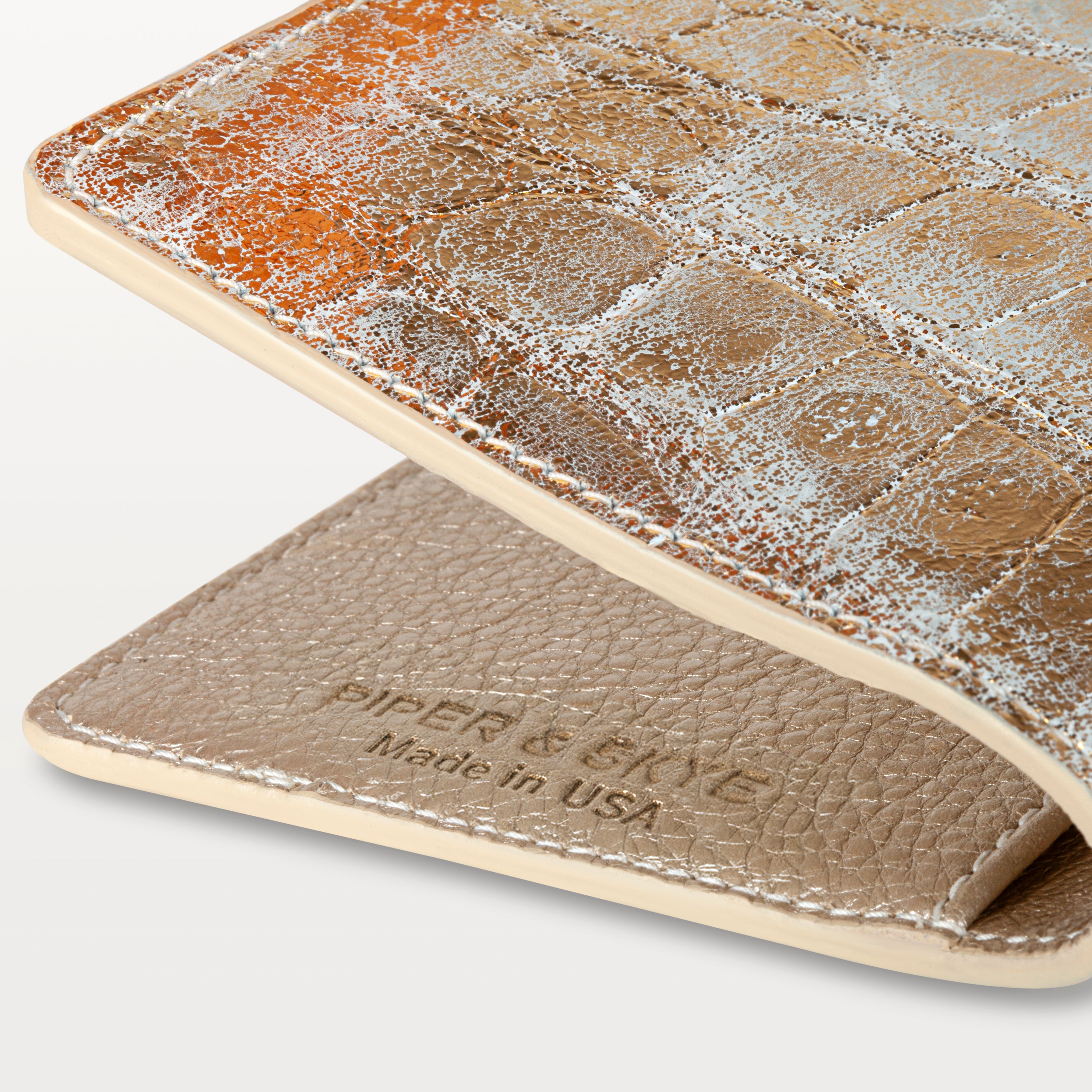 Close up view of the metallic gold alligator leather passport holder showing the textured pattern, embossed logo and 'Made in USA'. 