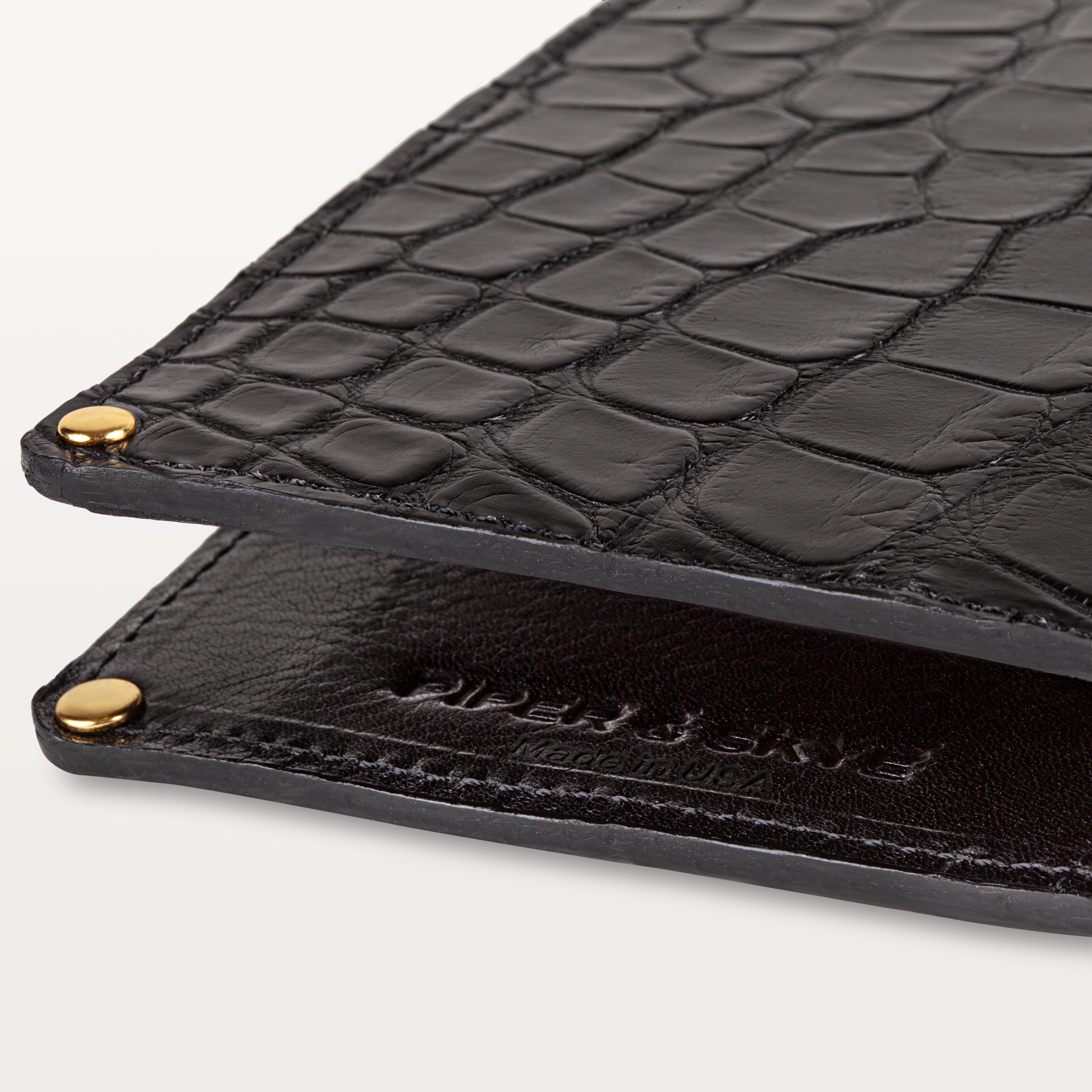 Close up view of the glossy black alligator leather passport holder showing the textured pattern, embossed logo and 'Made in USA'. 