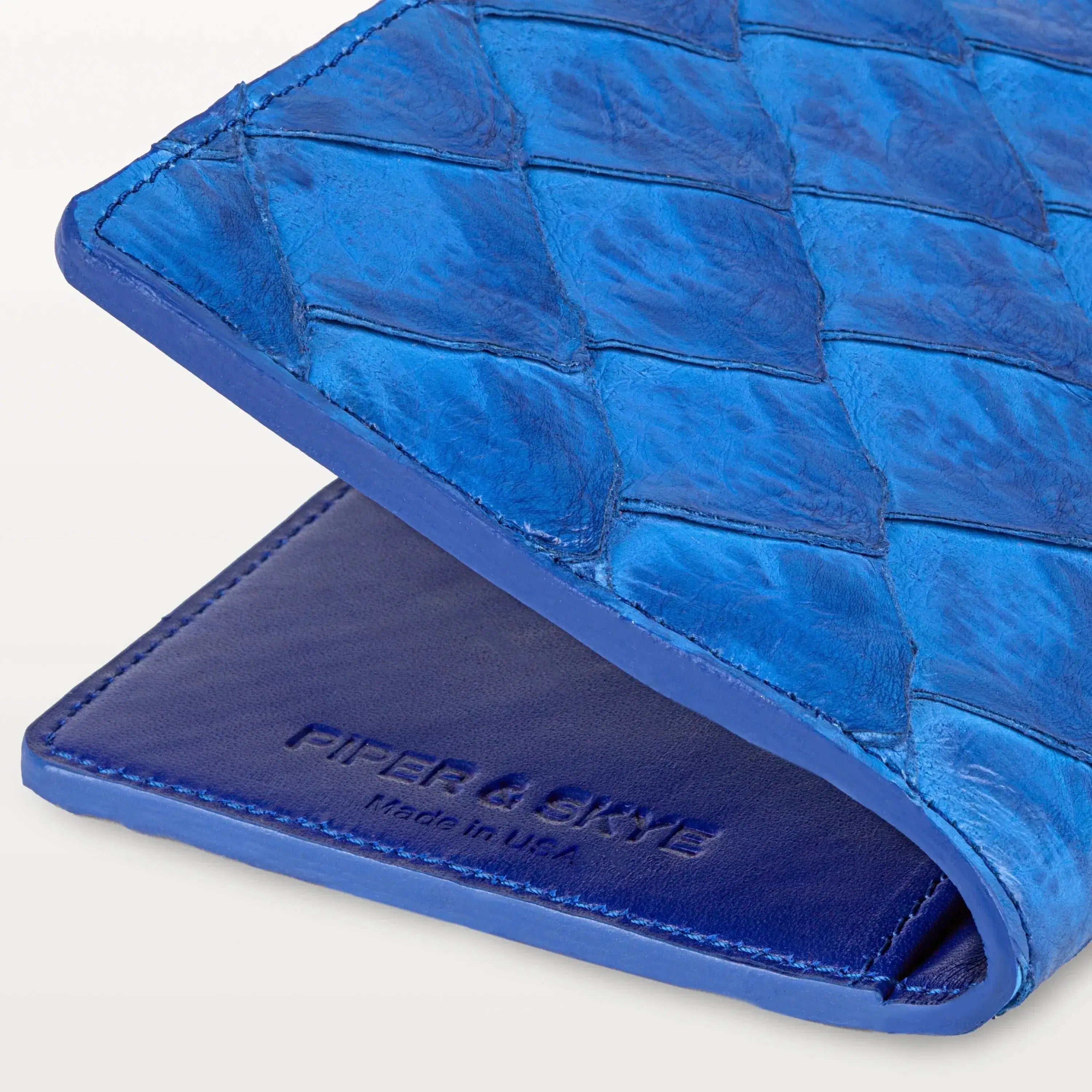 Close up view of the cobalt blue pirarucu passport holder showing the fish scale pattern, embossed logo and 'Made in USA'. 