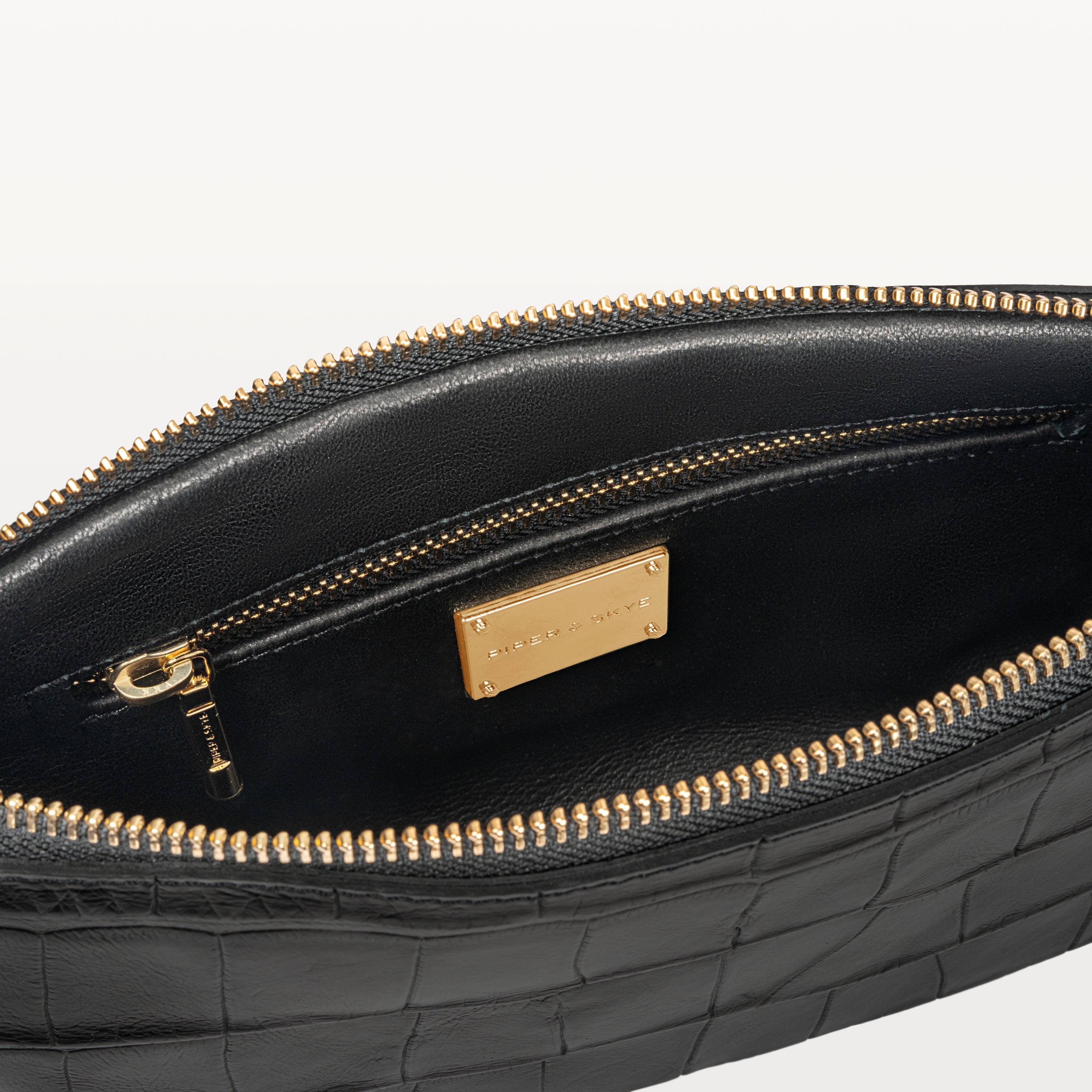 Interior view of the Nolita crossbody bag made from glossy black alligator, and its zippered inner pocket and gold Piper & Skye name plate. 