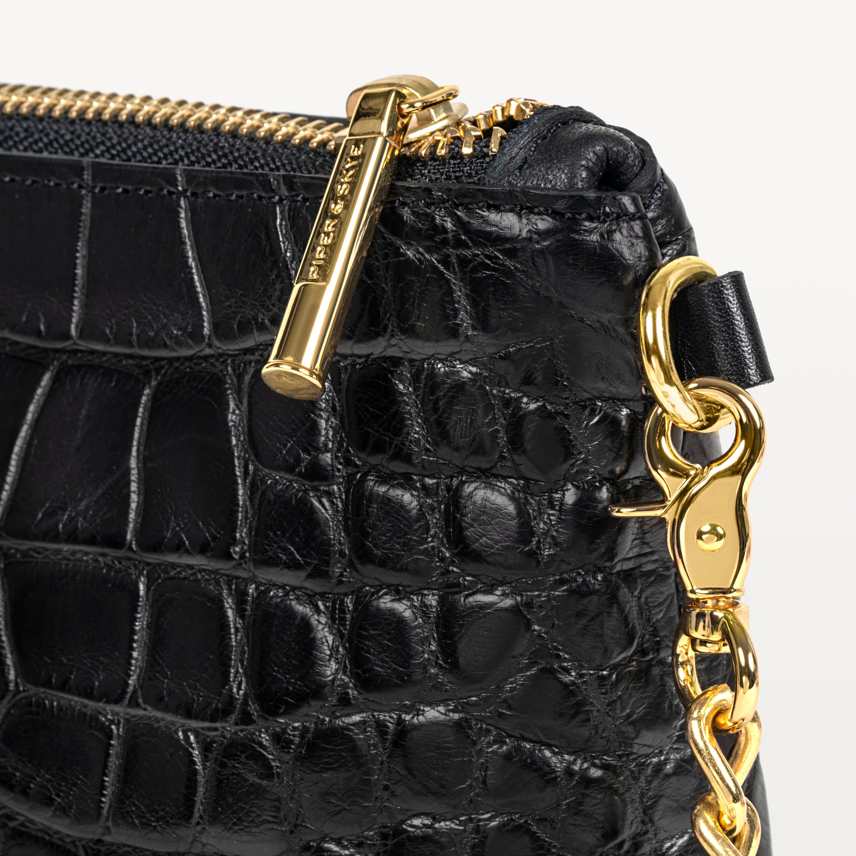 Close up view of the glossy black alligator material of the Nolita crossbody bag and it's gold zipper pull, D-ring, and crossbody strap. 